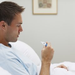 Mann schaut im Bett auf Fieberthermometer